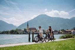 Two cyclists on a lakeside path with a village church and mountains in the background.