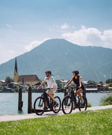 Two cyclists on a lakeside path with a village church and mountains in the background.