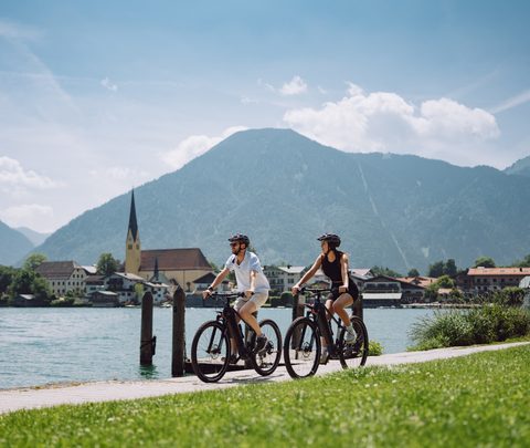 Two cyclists on a lakeside path with a village church and mountains in the background.