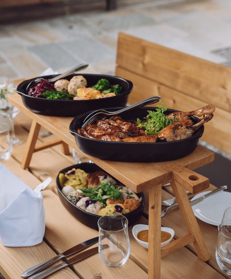 Warme Speisen in schwarzen Servierpfannen auf einem Holzgestell an einem gedeckten Restauranttisch.
