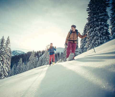 Two people hike with snowshoes across a snowy slope with fir trees and mountains in the background.