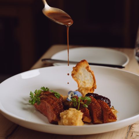 A plated meat dish on a white plate is being drizzled with sauce, with a glass of red wine beside it.
