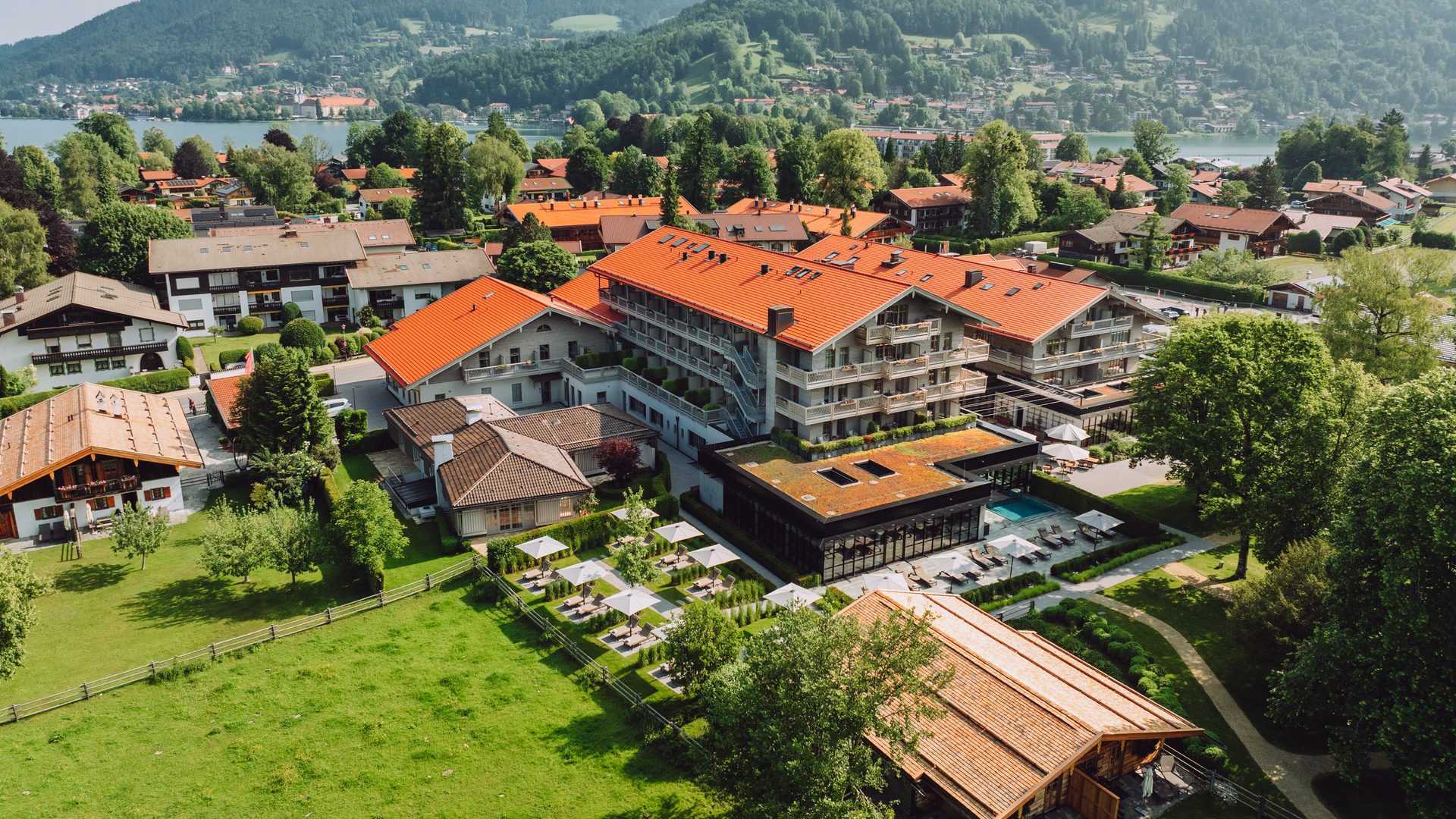 Aerial view of a hotel with pool, terrace and garden in a lakeside village with forested mountains.
