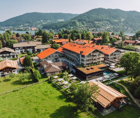 Luftaufnahme eines Hotels mit Pool, Terrasse und Garten in einem Dorf am See vor bewaldeten Bergen.