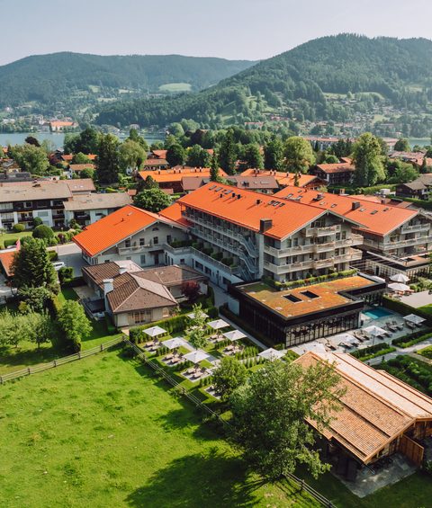 Luftaufnahme eines Hotels mit Pool, Terrasse und Garten in einem Dorf am See vor bewaldeten Bergen.