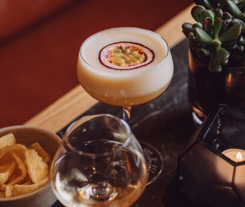 Cremiger Cocktail mit Fruchtgarnitur auf einem Tisch neben Glas, Snacks, Kerze und Pflanze.