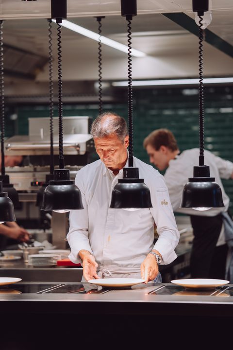 Chef plates dishes in an open restaurant kitchen while other staff work in the background.