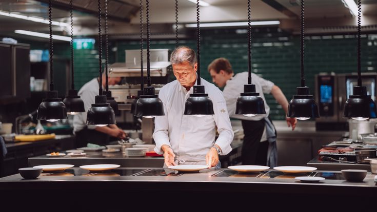 Koch richtet Teller in einer offenen Restaurantküche an, während weitere Mitarbeiter im Hintergrund arbeiten.