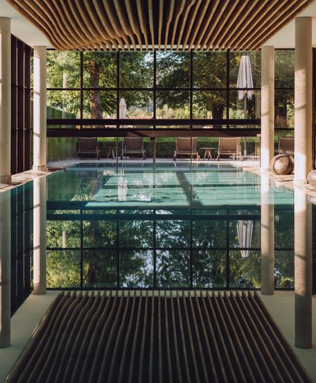 Innenpool mit großer Glasfront, Holzdecke und Liegen vor einem Garten.