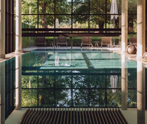 Innenpool mit großer Glasfront, Holzdecke und Liegen vor einem Garten.