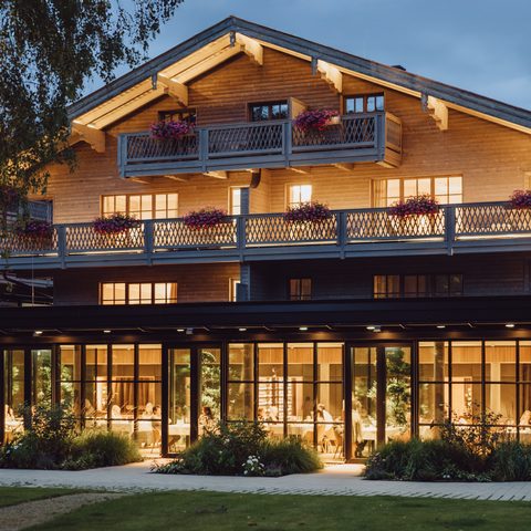 Mehrstöckiges Hotel im Chalet-Stil mit beleuchteter Glasfront, Balkonen und Garten in der Abenddämmerung.