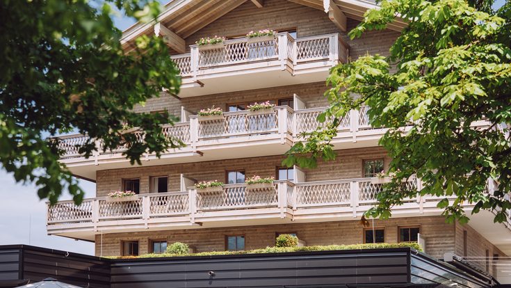 Mehrstöckiges Hotel im Chalet-Stil mit Holzbalkonen, Terrasse und Garten unter blauem Himmel.