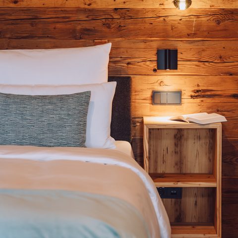 Bed with pillows beside a wooden bedside table and pendant light against a rustic wood wall.