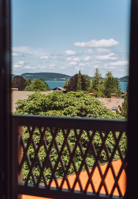 Blick durch ein Fenster über Bäume und Dächer auf einen See mit bewaldeten Hügeln im Hintergrund.