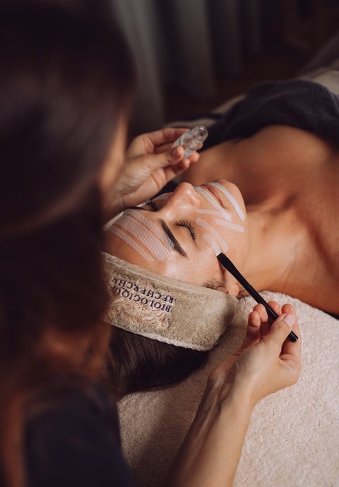 Kosmetikerin trägt bei einer Frau liegend im Spa eine Gesichtsmaske und Pflegeprodukt auf.