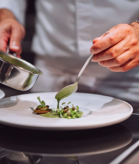 Ein Koch richtet ein Gericht auf einem weißen Teller an und gießt grüne Sauce darüber.