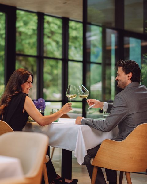 A couple sits in a restaurant and toasts with two white wine glasses at a set table.
