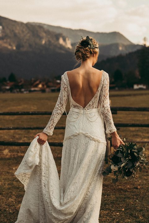 Bride in a long lace dress with bouquet on a meadow in front of mountains.