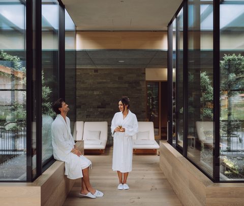 Zwei Gäste im Bademantel in einem modernen Wellness-Ruhebereich mit Glasfronten und Liegen.