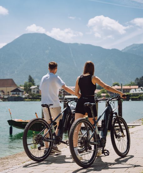A couple with bicycles stands by the lakeshore in front of mountains and a village with a church steeple.