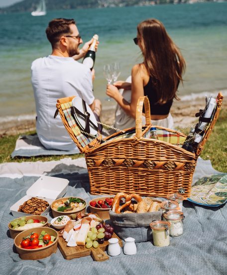 A couple sits at a lakeside picnic with a basket, snacks and a bottle of sparkling wine.