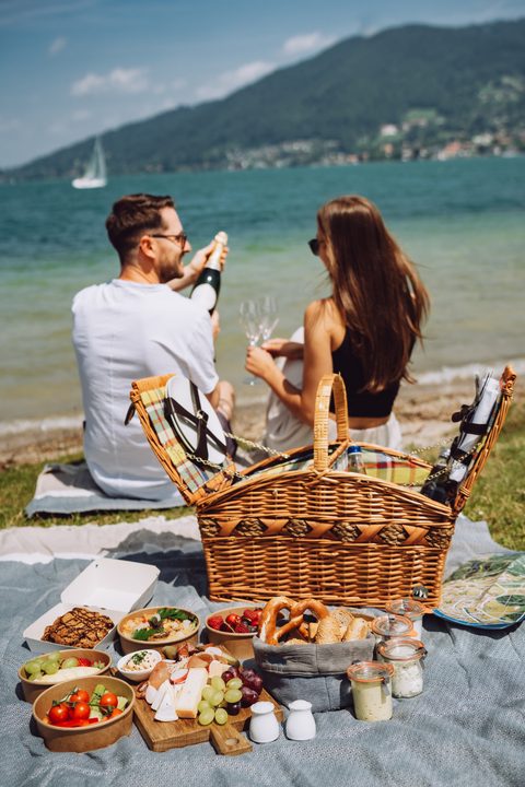 A couple sits at a lakeside picnic with a basket, snacks and a bottle of sparkling wine.