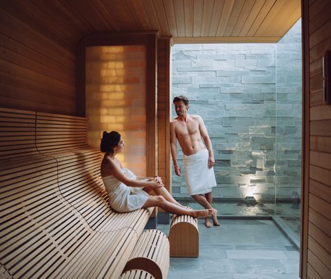 Ein Paar mit Handtüchern entspannt in einer Sauna mit Holzbank und Glaswand zum angrenzenden Spa-Bereich.