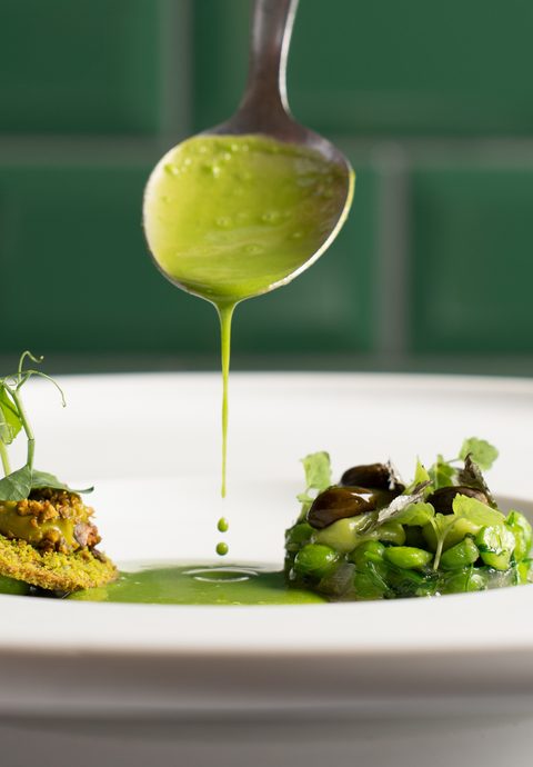 Ein angerichtetes Gourmetgericht mit Gemüse und grüner Sauce auf einem weißen Teller, darüber ein Löffel mit Sauce.