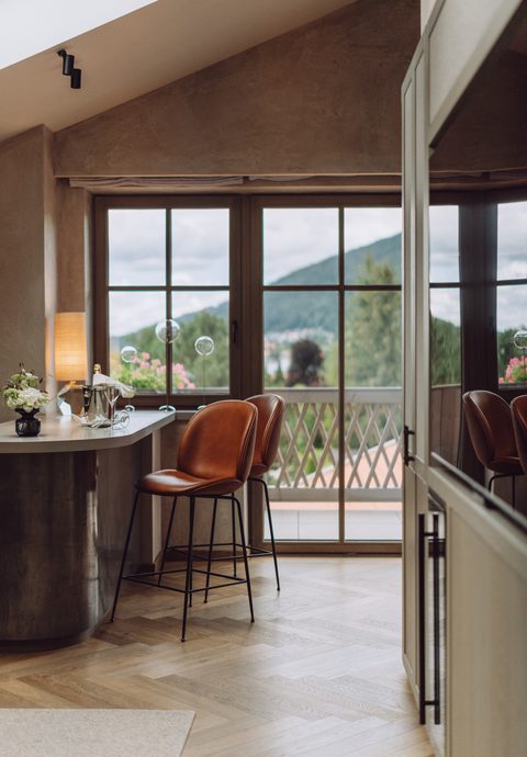 Sitzbereich in einem Hotelzimmer mit Hochtisch, zwei Lederstühlen und Balkontür mit Bergblick.