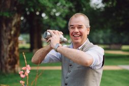 Ein Barkeeper mixt im Garten mit einem Cocktailshaker Getränke an einem Tisch mit Gläsern und Blumen.