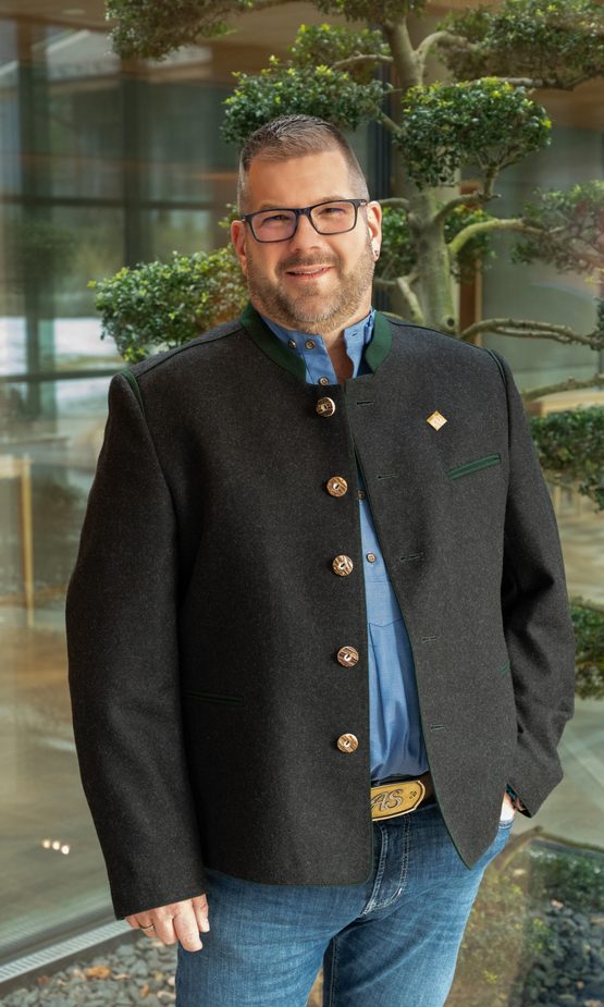 Lächelnder Mann mit Brille in Trachtenjacke steht in einem Hotelinnenbereich vor Glasfront und Bonsai.