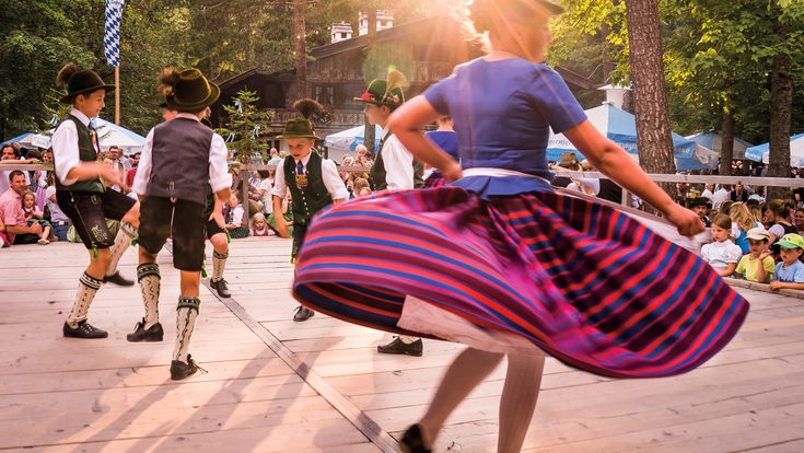 Kinder und Jugendliche tanzen in bayerischer Tracht auf einer Freiluftbühne vor Publikum im Wald.
