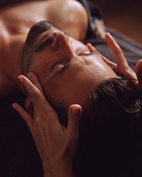 Ein Mann liegt bei einer Gesichts- und Kopfmassage im Wellnessbereich mit geschlossenen Augen.