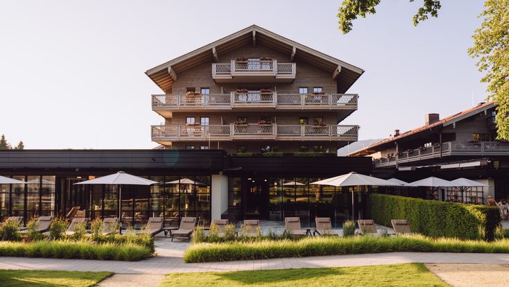 Außenansicht eines Hotels mit Holzfassade, Balkonen, Terrasse, Liegestühlen und Garten.