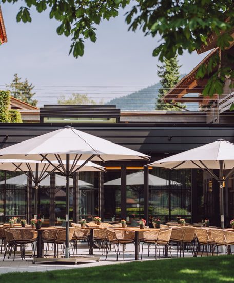 Restaurantterrasse mit gedeckten Tischen, Korbstühlen und Sonnenschirmen in einem grünen Hotelinnenhof.