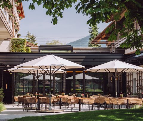 Restaurantterrasse mit gedeckten Tischen, Korbstühlen und Sonnenschirmen in einem grünen Hotelinnenhof.