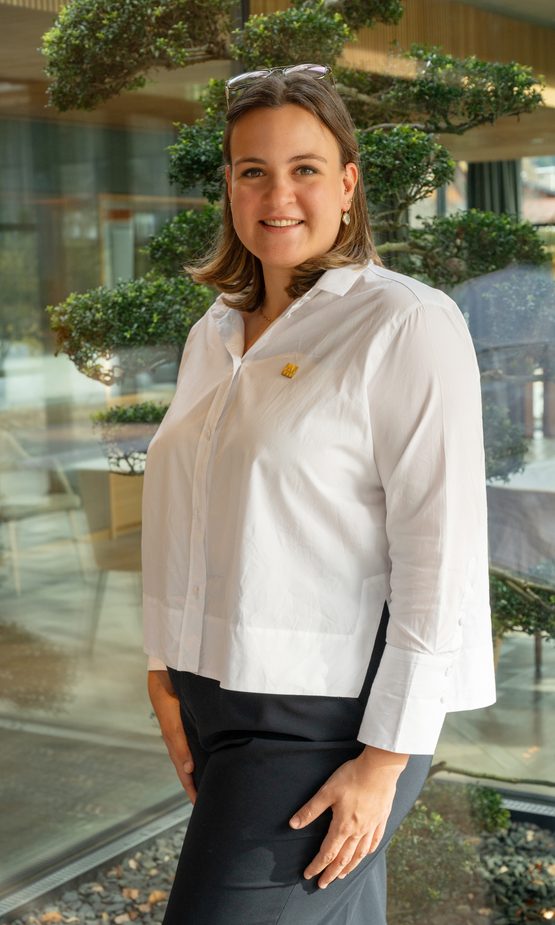 Hotelmitarbeiterin in weißer Bluse steht in der Lobby vor einer Bonsai-Pflanze und blickt in die Kamera.
