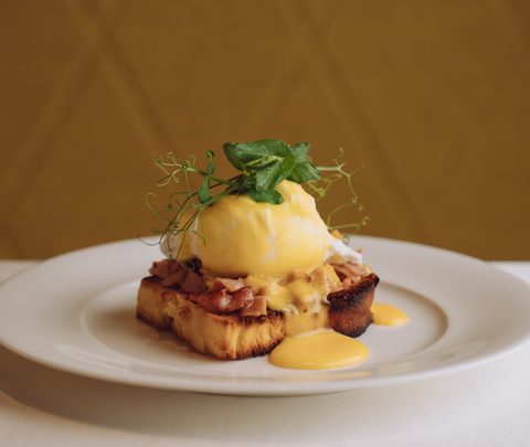 Eggs Benedict mit pochiertem Ei, Sauce Hollandaise, Speck und Kräutern auf einem weißen Teller.
