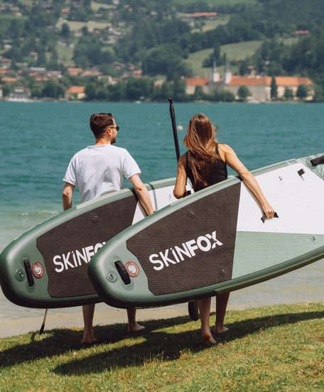 Two people carry stand-up paddle boards along the shore of a lake.