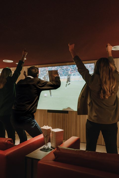 Drei Gäste jubeln vor einer großen Projektion eines Fußballspiels in einem gemütlichen Innenraum mit Sesseln und Popcorn.