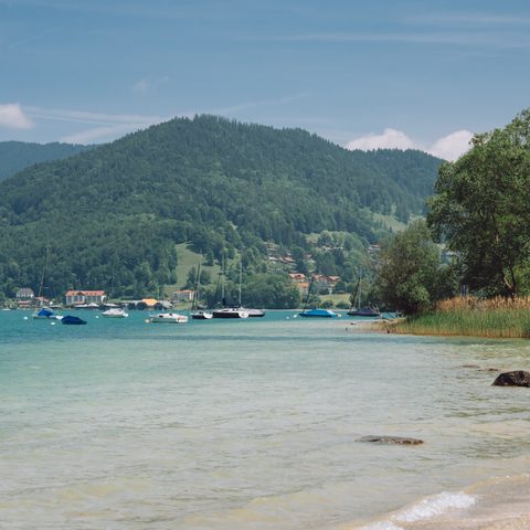 Lakeshore with shallow water, several boats, and forested mountains in the background.