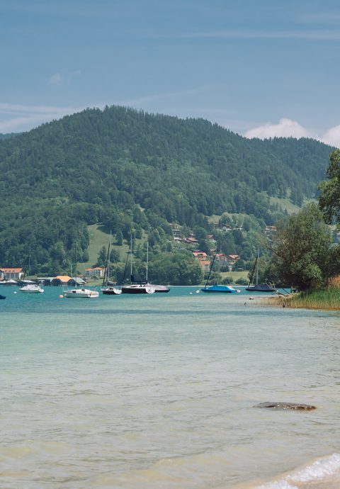 Lakeshore with shallow water, several boats, and forested mountains in the background.