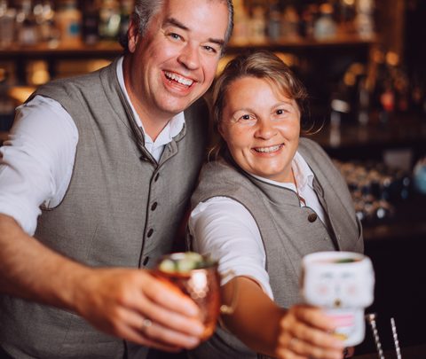 Zwei Barkeeper hinter einer Bar halten Getränke in die Kamera und lächeln.