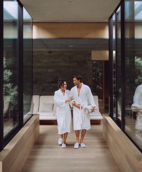 Ein Paar in Bademänteln geht durch einen modernen Wellnessbereich mit Glaswänden und Sitzbank.