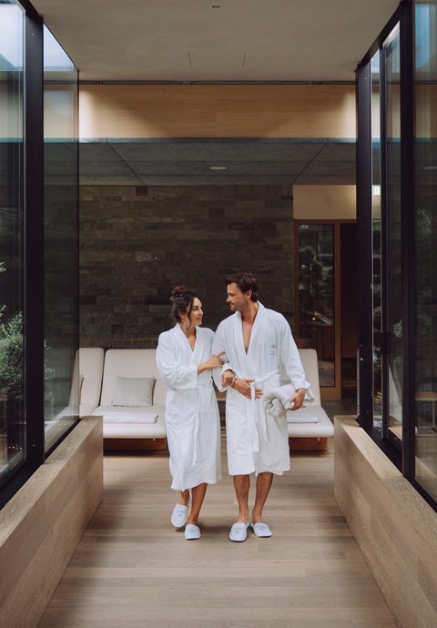 A couple in bathrobes walks through a modern wellness area with glass walls and a bench.