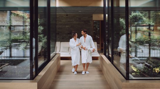 A couple in bathrobes walks through a modern wellness area with glass walls and a bench.