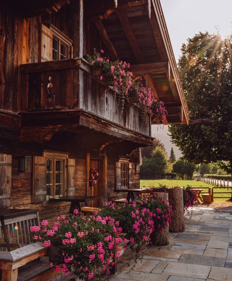 Traditionelles Holzchalet mit Balkon, pinken Blumenkästen und gepflasterter Terrasse im Außenbereich.