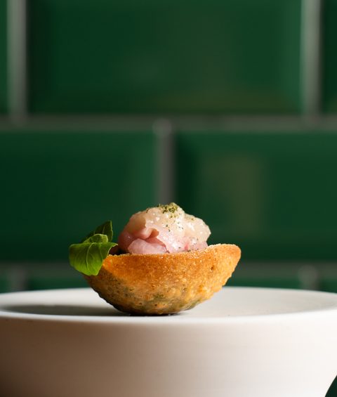 Small appetizer in a crisp shell with tartare and lettuce leaf on white plate in front of green tiles.