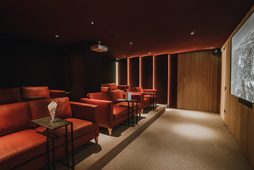 Abgedunkelter Kinoraum mit roten Sesseln, Beistelltischen, Projektor und großer Leinwand.