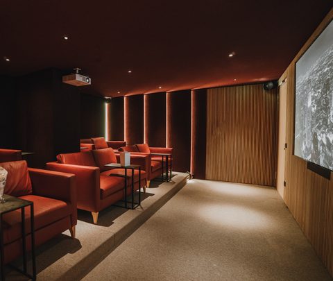 Darkened cinema room with red armchairs, side tables, projector and large screen.